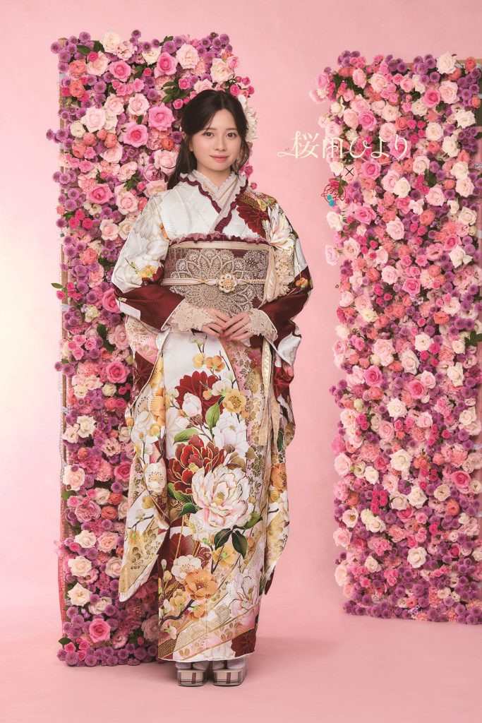 桜田ひより着用の白地振袖。赤茶色を基調に牡丹や花柄が華やかに描かれた成人式向け振袖。ピンクの花装飾背景で上品かつ可憐な振袖コーディネート。