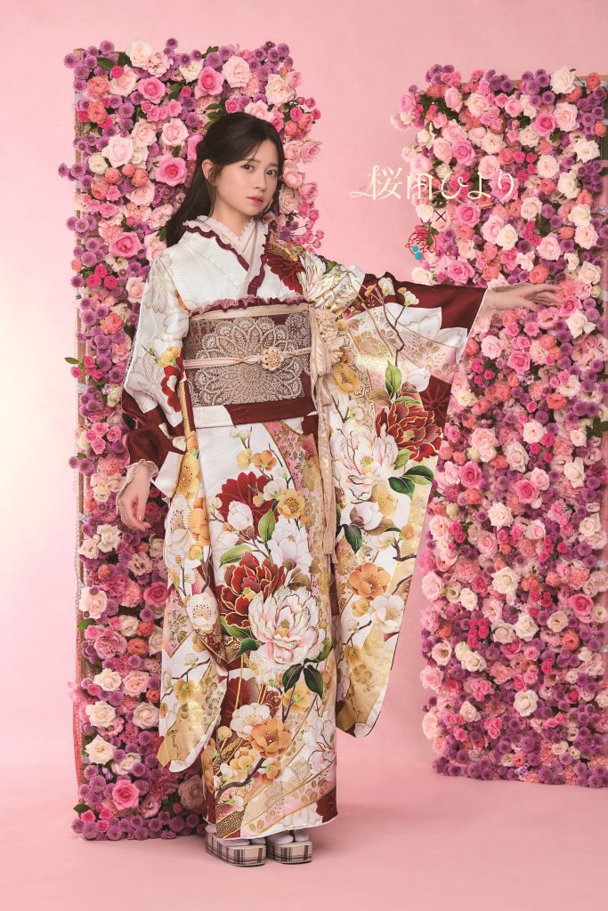 桜田ひより着用の白地振袖。赤茶色を基調に牡丹や花柄が華やかに描かれた成人式向け振袖。ピンクの花装飾背景で上品かつ可憐な振袖コーディネート。