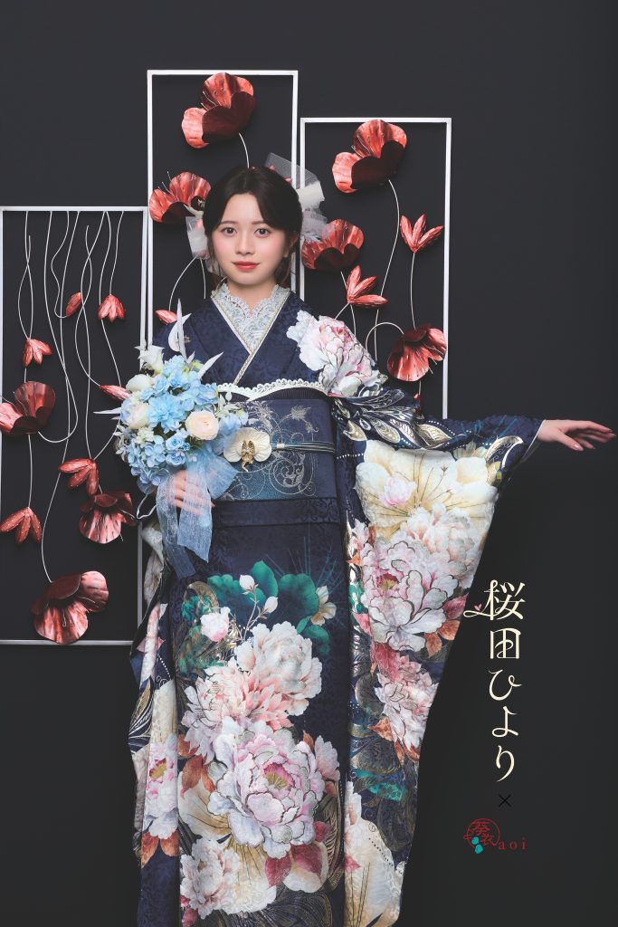 桜田ひより着用の紺地振袖。ネイビーを基調に大輪の牡丹や花柄が描かれた、シックで華やかな成人式向け振袖。黒背景とアートフラワー装飾で写真映えする振袖コーディネート。