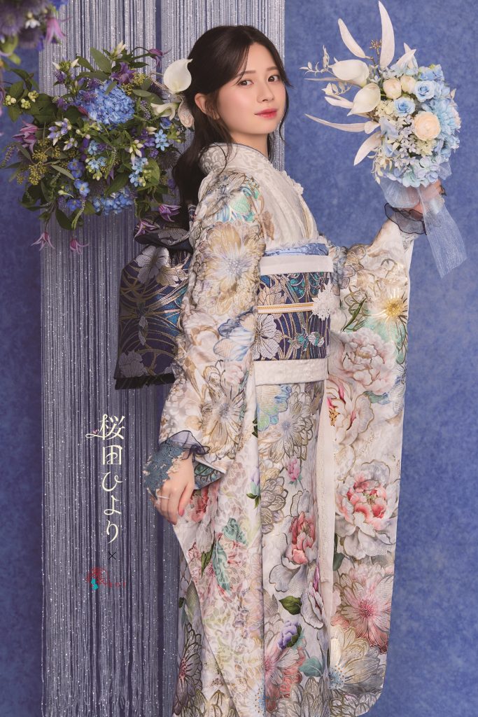 桜田ひより着用の白地×赤茶色振袖。大輪の牡丹と花柄が華やかに描かれた成人式向け振袖スタイル。ピンクの花装飾背景で上品かつ可憐な雰囲気の振袖コーディネート。