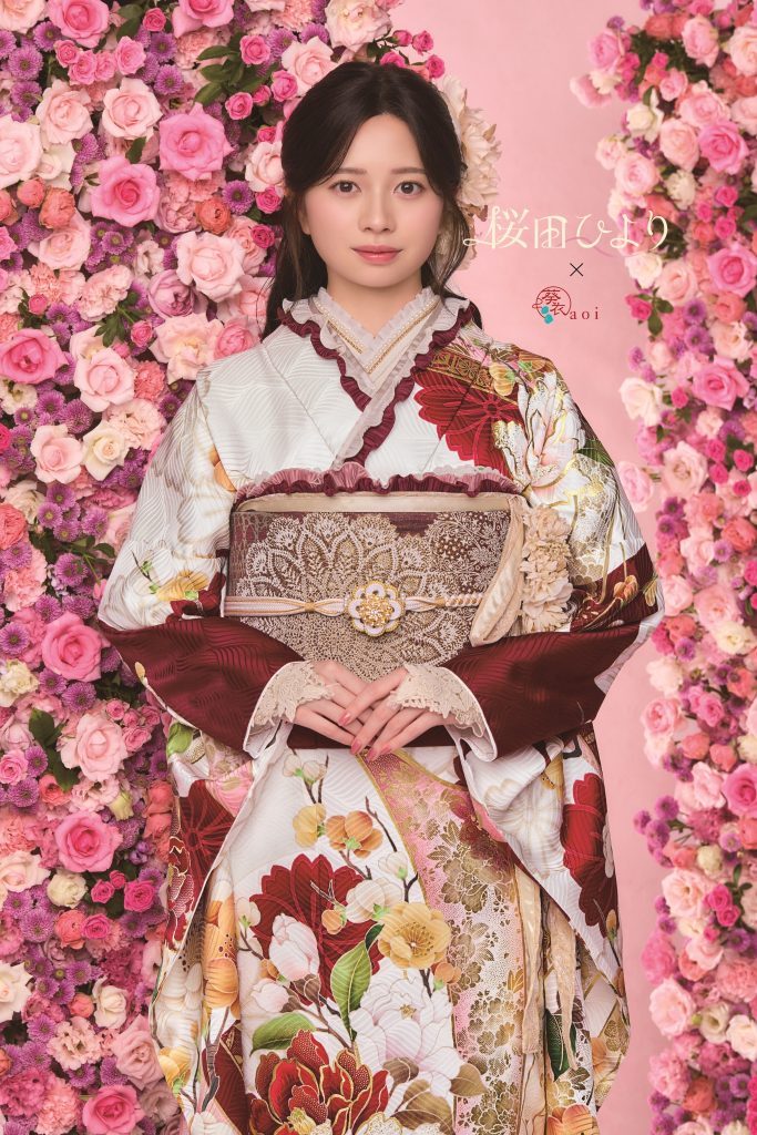 桜田ひより着用の白地振袖。赤茶色を基調に牡丹や花柄が華やかに描かれた成人式向け振袖。ピンクの花装飾背景で上品かつ可憐な振袖コーディネート。