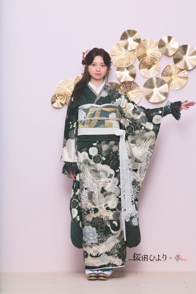 桜田ひより着用の深緑系振袖。松や鶴などの古典柄と花文様が描かれた、格式と華やかさを兼ね備えた成人式向け振袖。ゴールド装飾背景で上品かつモダンな振袖コーディネート。